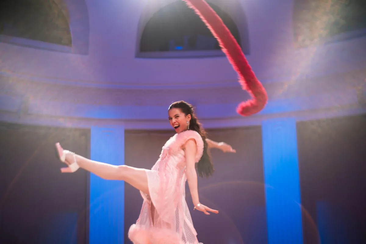 A performer in a pale pink costume mid-movement under stage lighting
