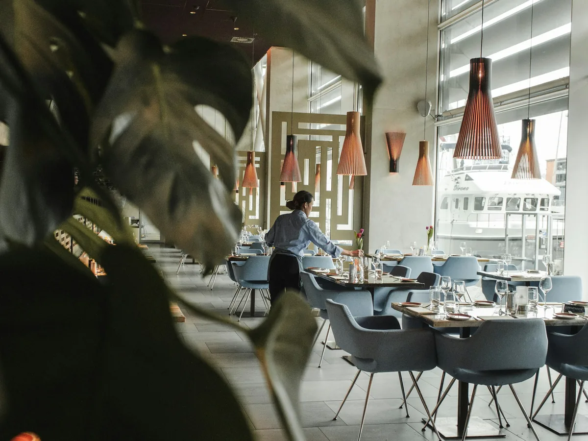 A restaurant dining room set with tables, hanging pendant lights and tall windows